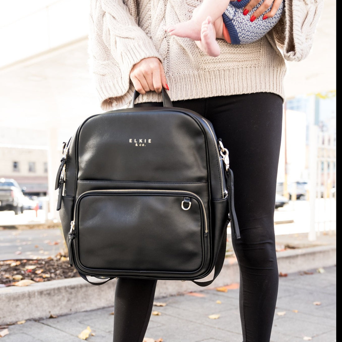 woman in the city holding a baby and also holding a black vegan leather backpack by  the  top grab handle