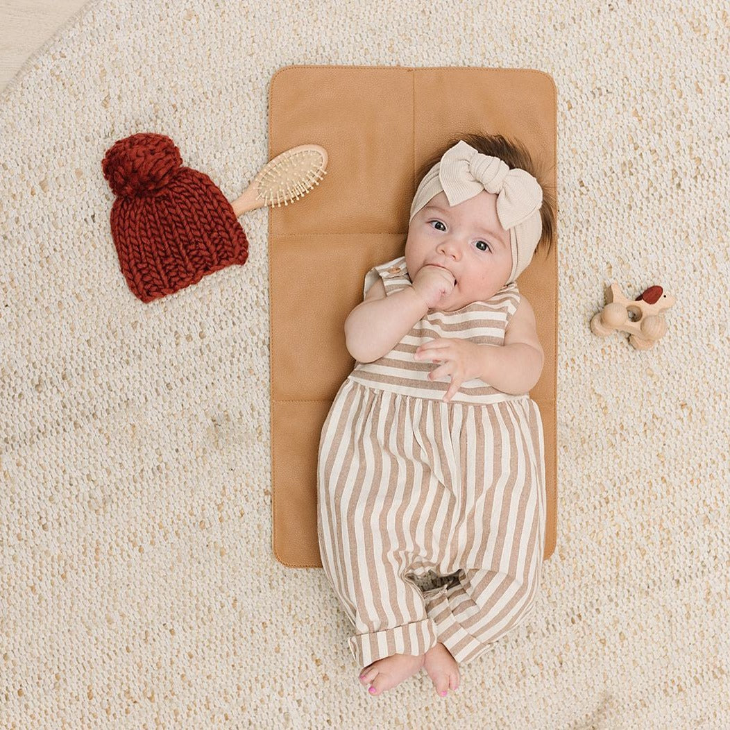 baby on Camel vegan leather changing mat with a tan and white stripe romper and a bow headband. Baby has her fist in her mouth. There is a red crocheted beanie hat with pom, fephus wooden hair brush and wooden dog toy nearby. The rug is a braided cream color.